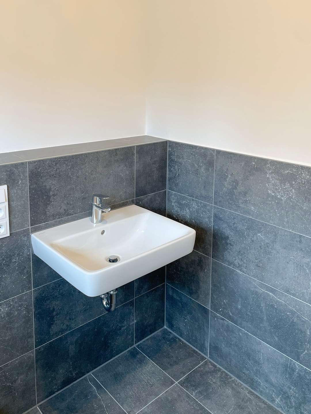 White wall-mounted sink with chrome faucet in a corner bathroom with dark gray tiled walls and floor.