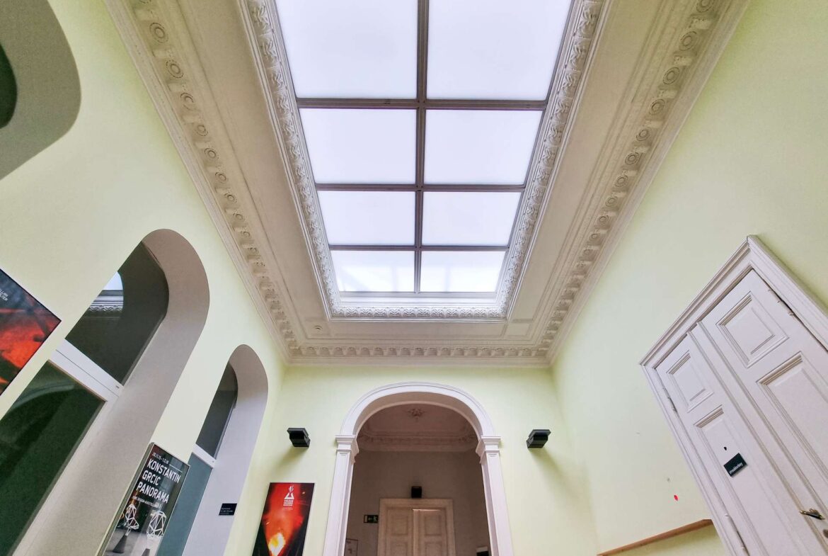 Interior of a pale-green hallway with a large central skylight and ornate moldings along the ceiling.