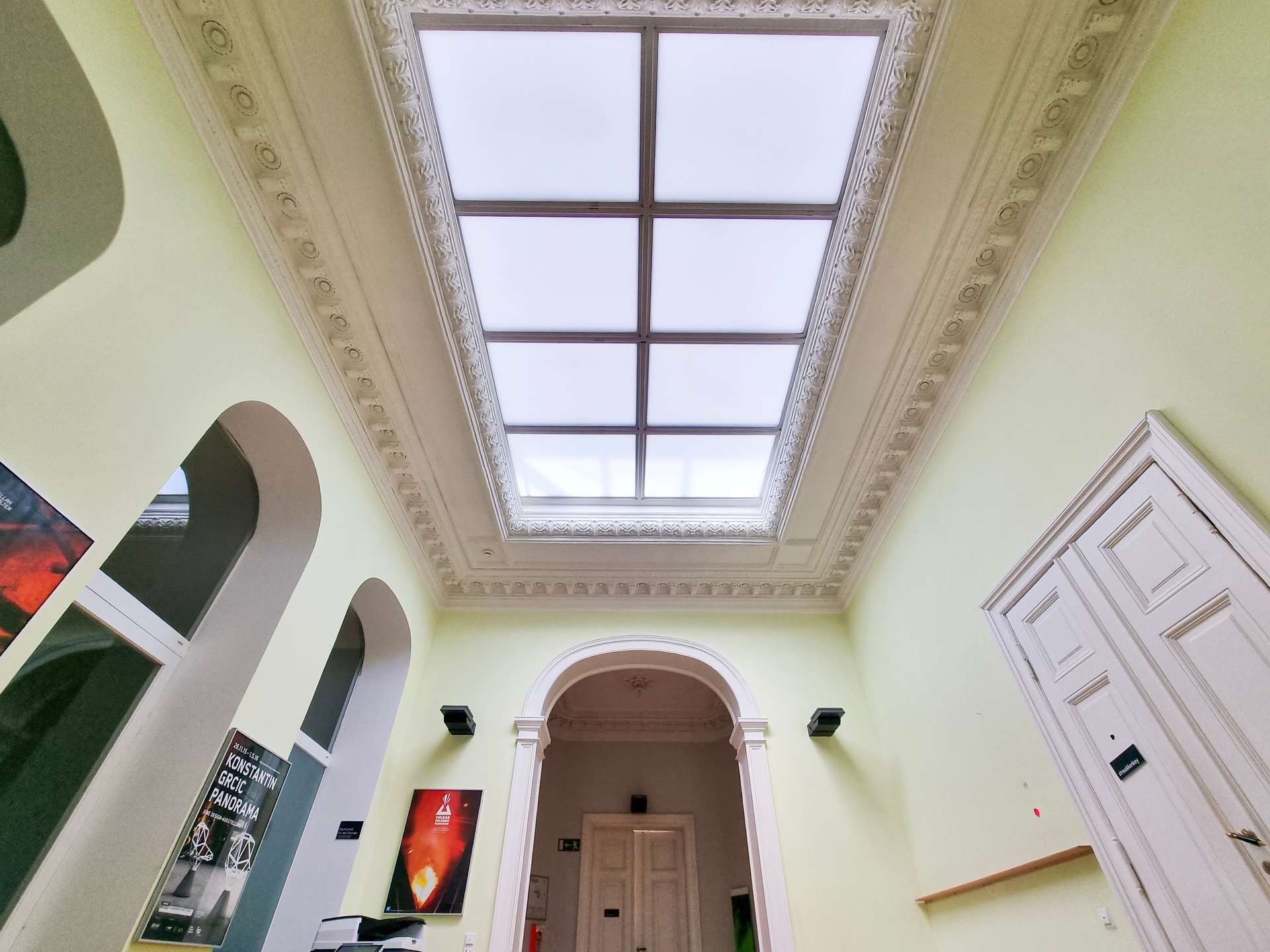 Interior of a pale-green hallway with a large central skylight and ornate moldings along the ceiling.