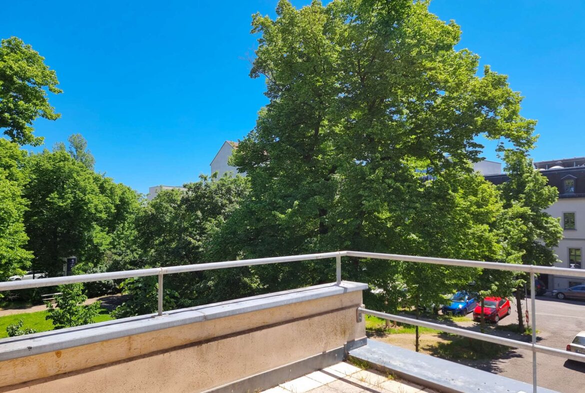 Balcony railing and concrete ledge with lush trees and a bright blue sky beyond, in an urban setting