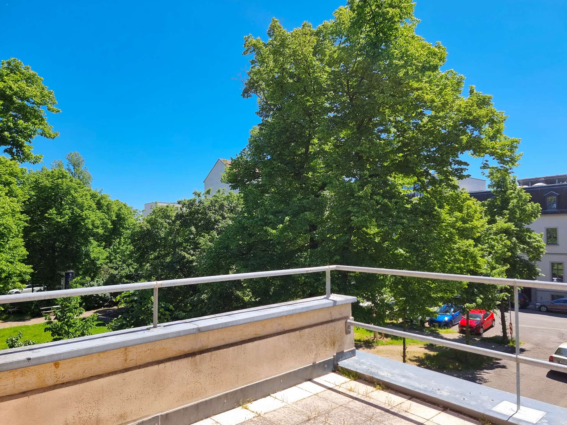Balcony railing and concrete ledge with lush trees and a bright blue sky beyond, in an urban setting