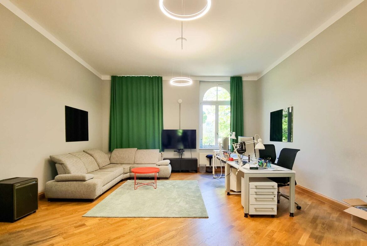 Living room with a gray sectional sofa, green curtains, and a desk area on the right under round LED ceiling lights.