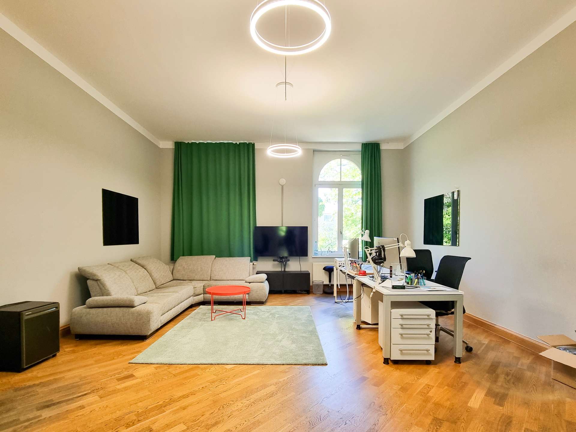 Living room with a gray sectional sofa, green curtains, and a desk area on the right under round LED ceiling lights.