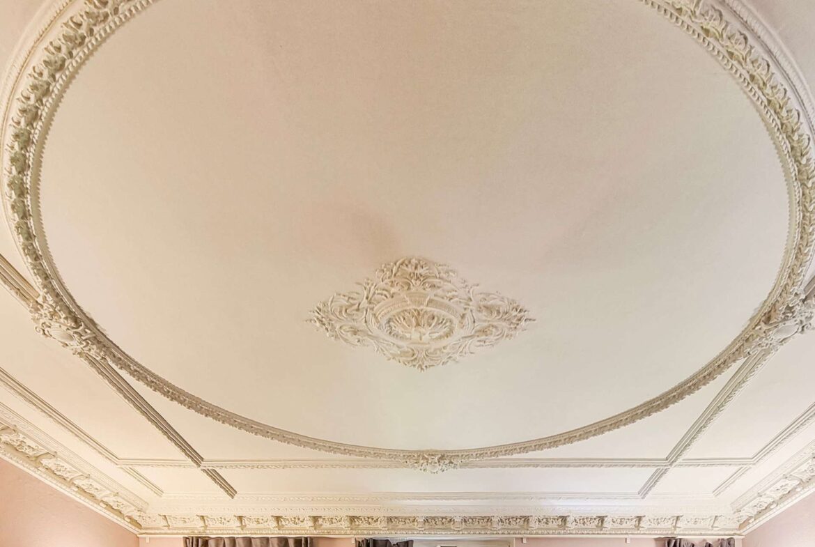 Ornate plaster ceiling with a central decorative medallion and detailed molding, seen above draped windows in a beige room