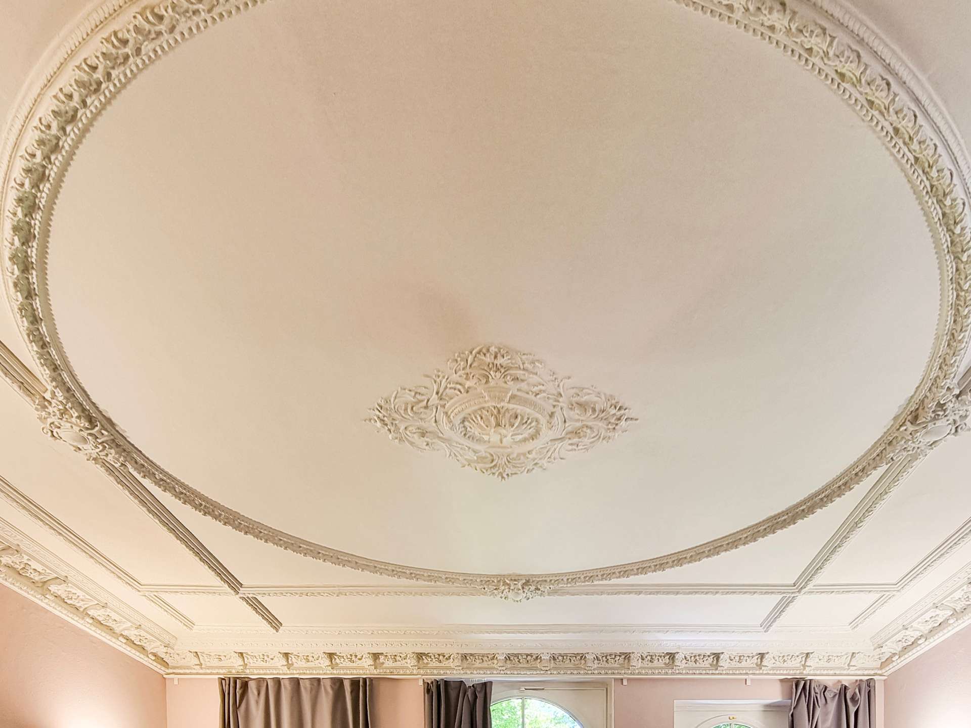 Ornate plaster ceiling with a central decorative medallion and detailed molding, seen above draped windows in a beige room