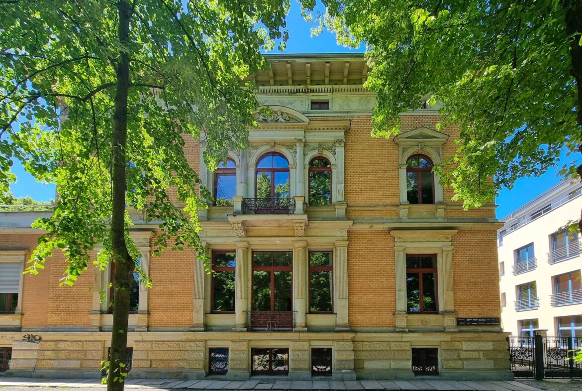 Historic brick building with ornate stone arches and tall arched windows, framed by green trees on a sunny day.