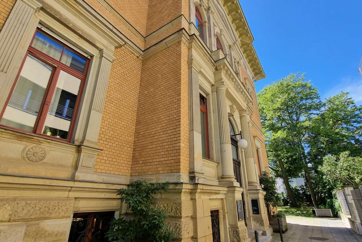 Corner of an ornate beige brick building with decorative columns, red-framed windows, and a bright blue sky.