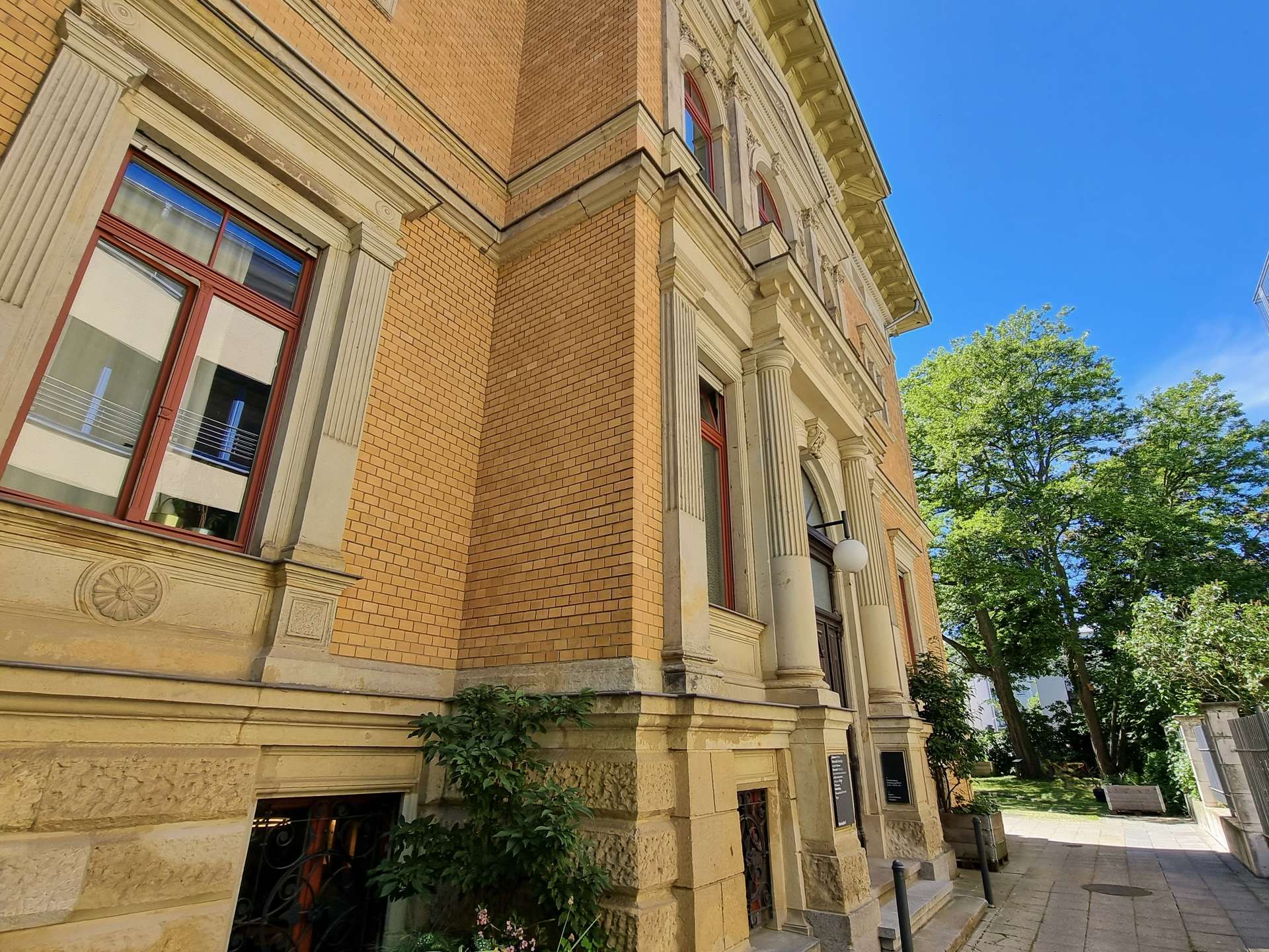 Corner of an ornate beige brick building with decorative columns, red-framed windows, and a bright blue sky.