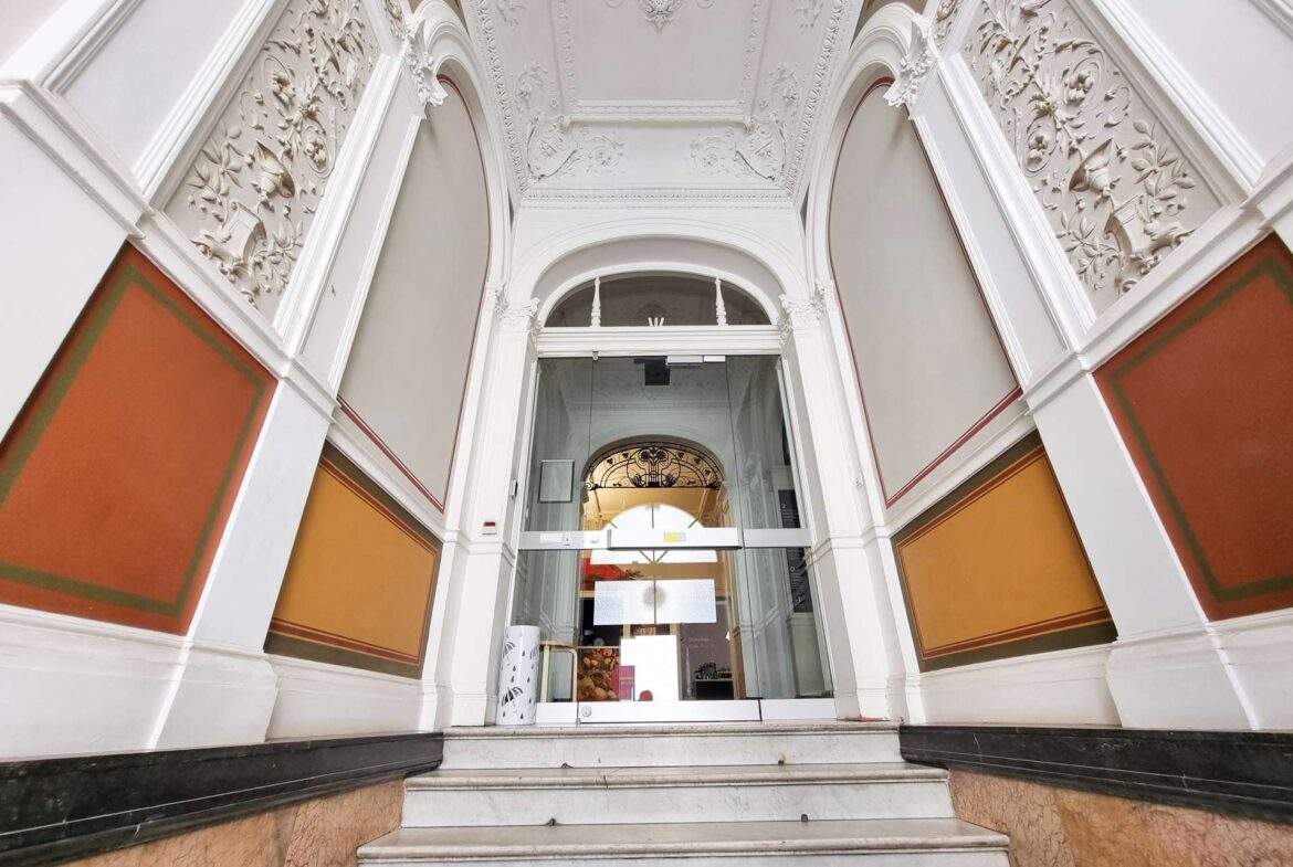 Ornate white facade with intricate plasterwork around a glass doorway, flanked by orange wall panels and marble steps.
