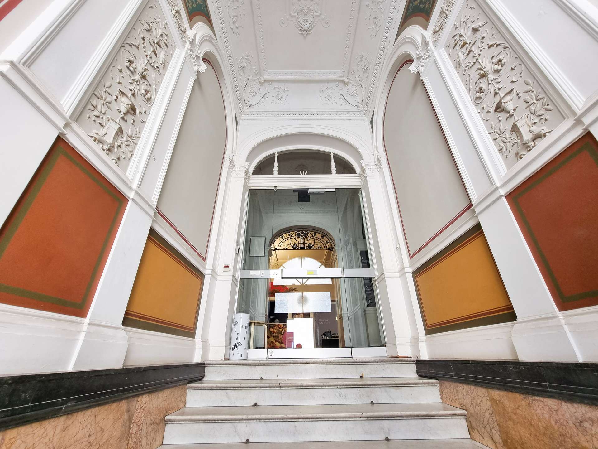 Ornate white facade with intricate plasterwork around a glass doorway, flanked by orange wall panels and marble steps.