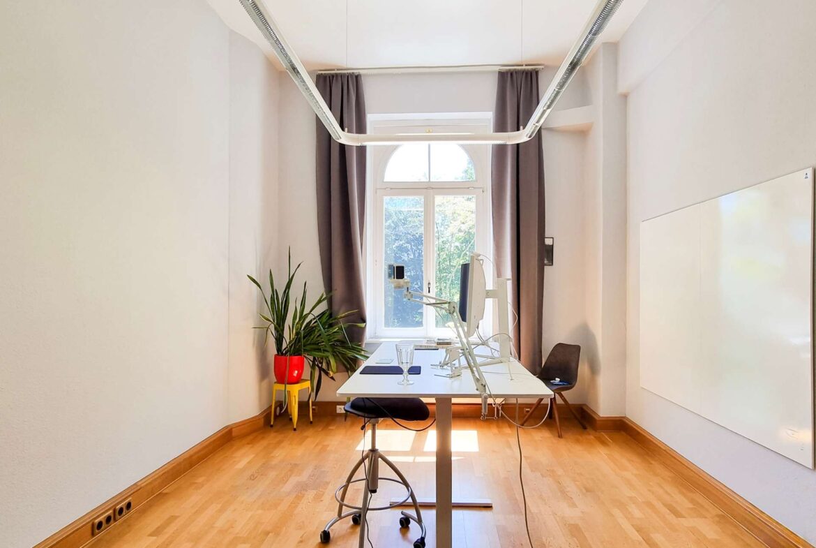 Bright office/workstation with a long white desk, monitor arms, and a window with curtains in a sunlit room.