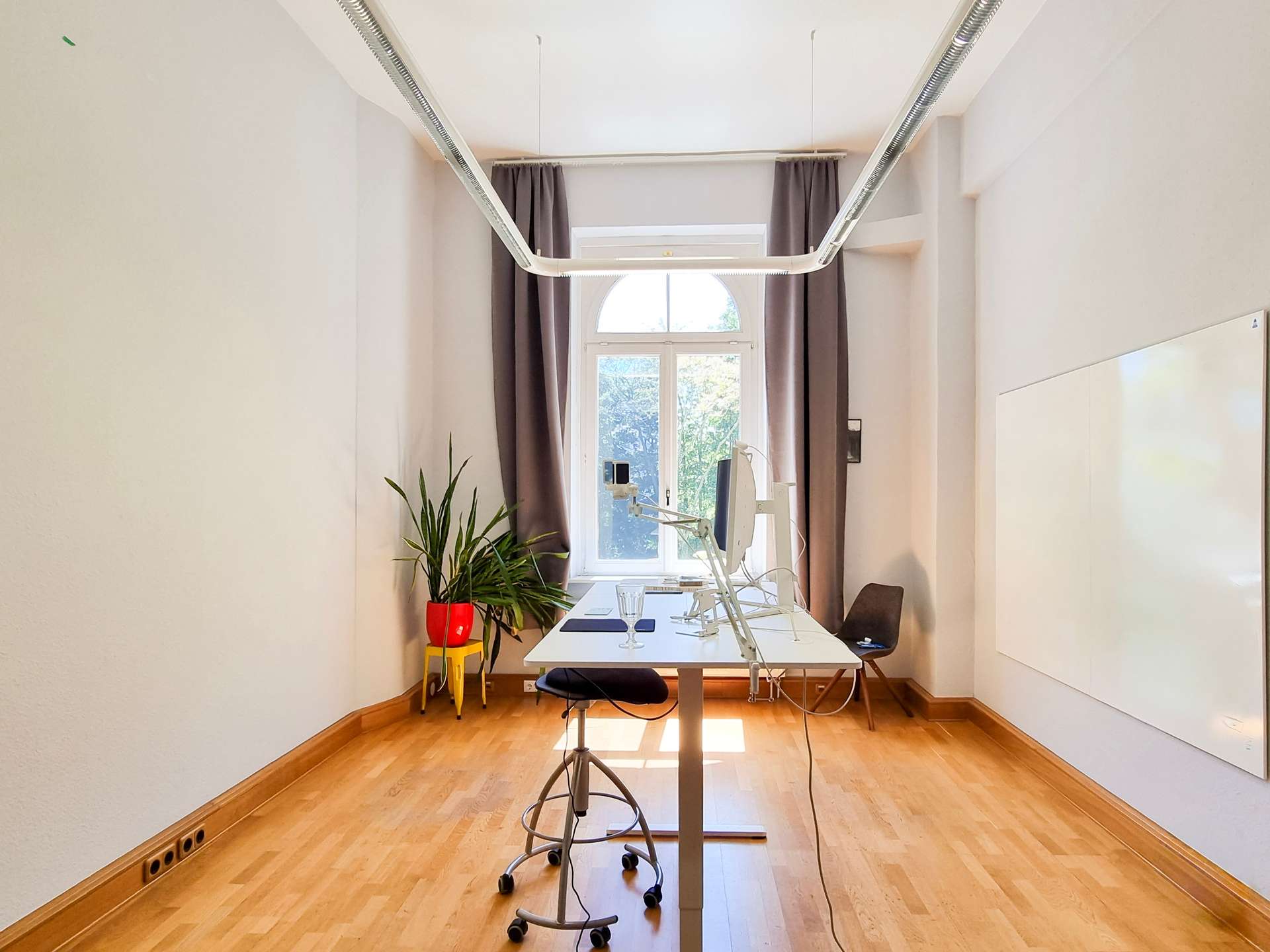 Bright office/workstation with a long white desk, monitor arms, and a window with curtains in a sunlit room.