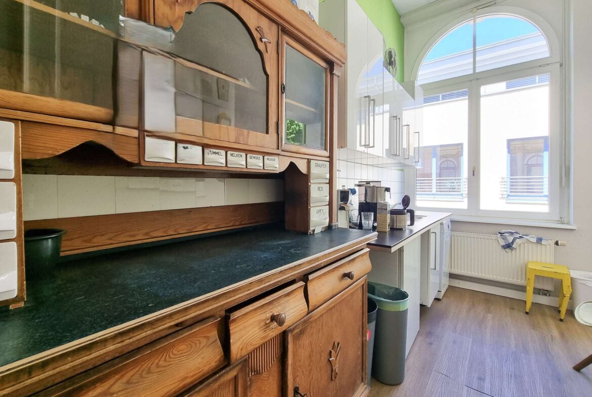Antique wooden kitchen cabinet and counter inside a bright green-walled cafe, with a large arched window on the right.