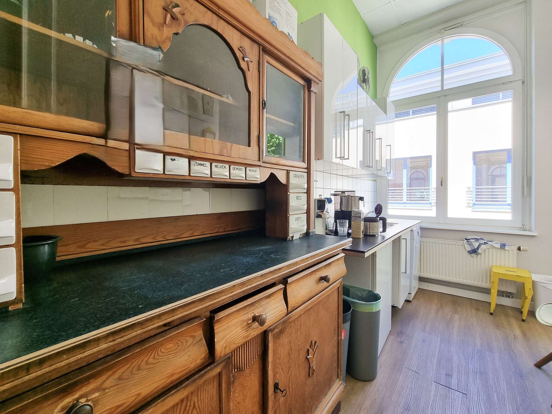 Antique wooden kitchen cabinet and counter inside a bright green-walled cafe, with a large arched window on the right.