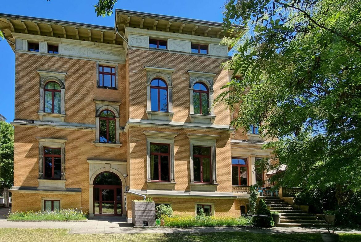 Brick multi-story building with arched windows surrounded by stone frames, set beside a green garden and blue sky framed by trees