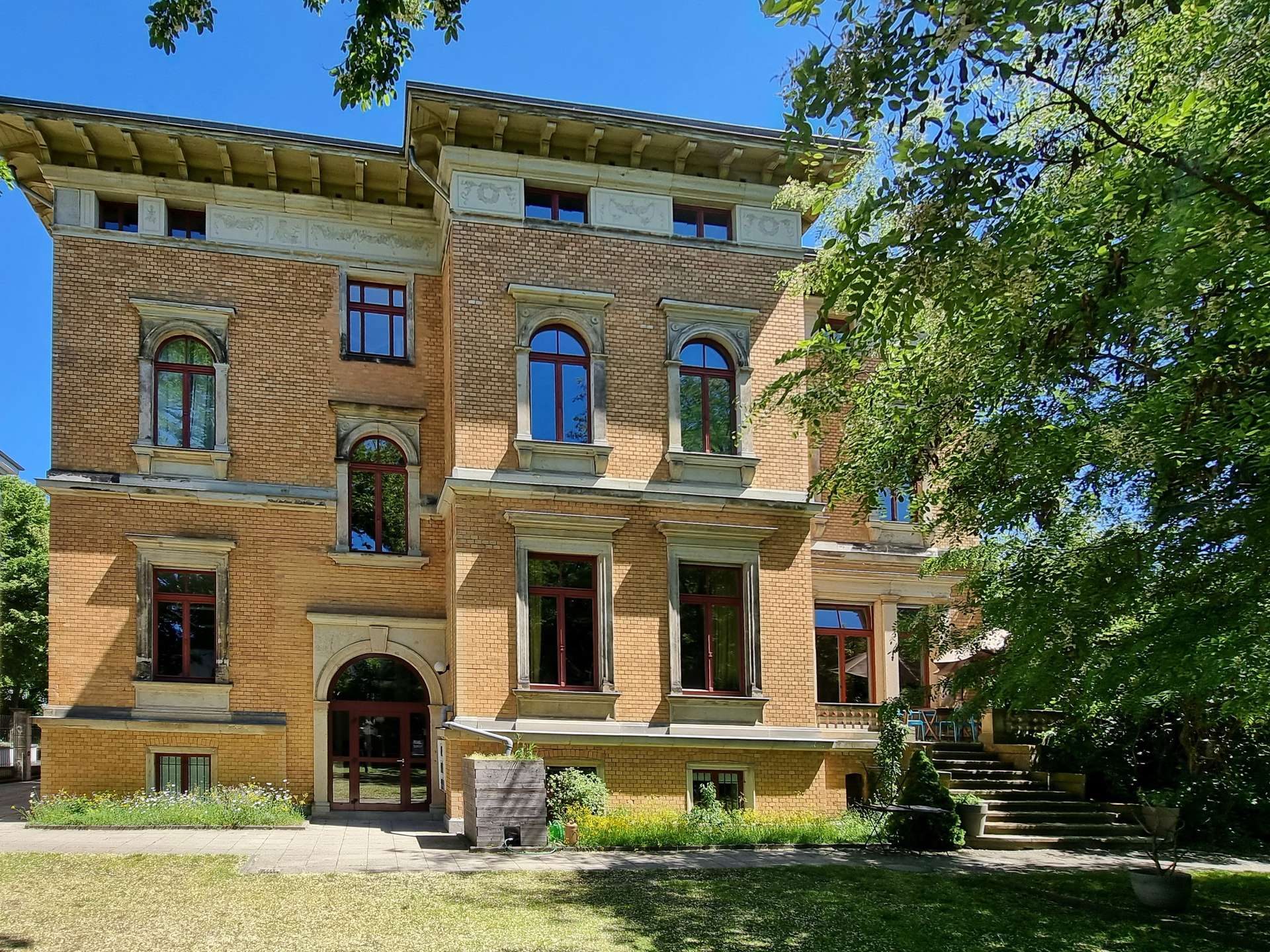 Brick multi-story building with arched windows surrounded by stone frames, set beside a green garden and blue sky framed by trees