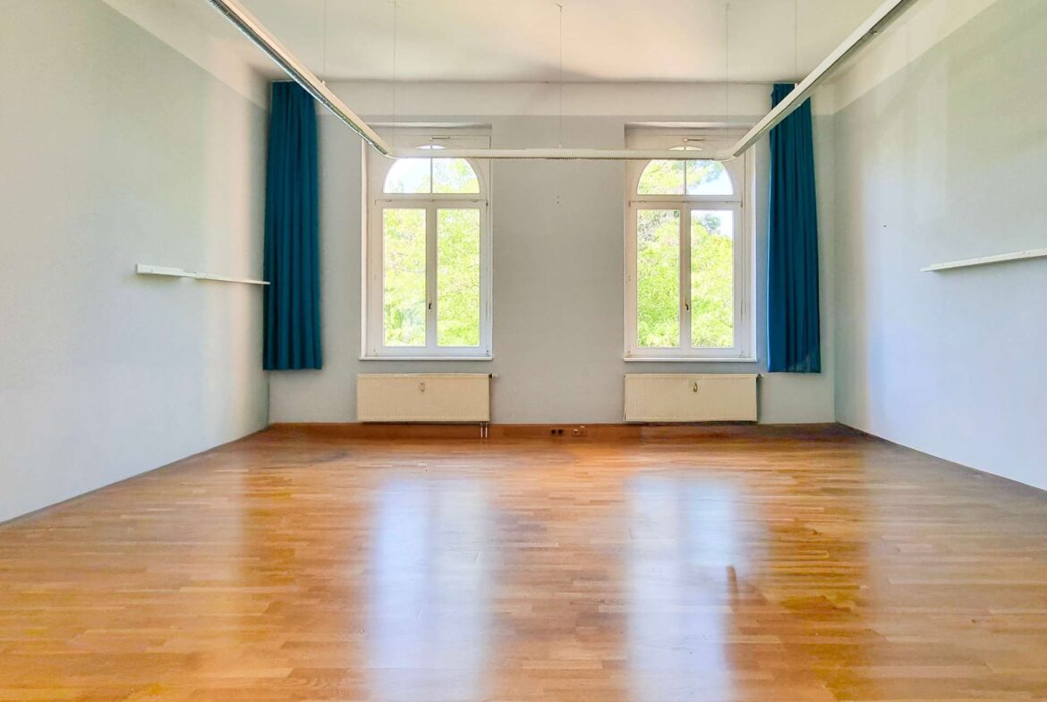 Empty dance/studio room with a wooden floor, pale blue walls, and blue curtains beside two large arched windows letting in greenery.