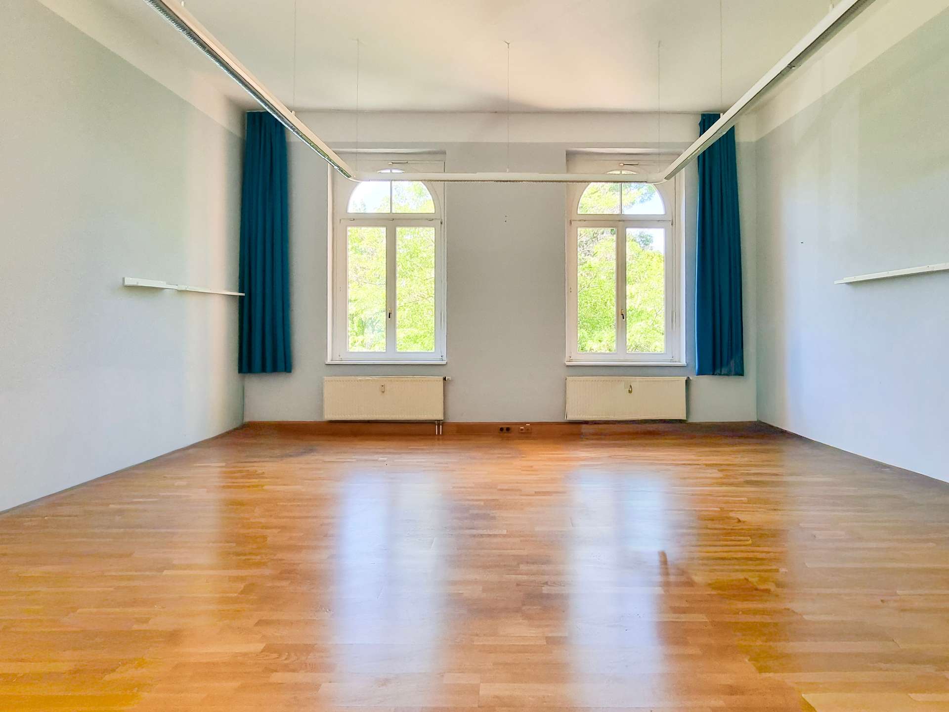 Empty dance/studio room with a wooden floor, pale blue walls, and blue curtains beside two large arched windows letting in greenery.
