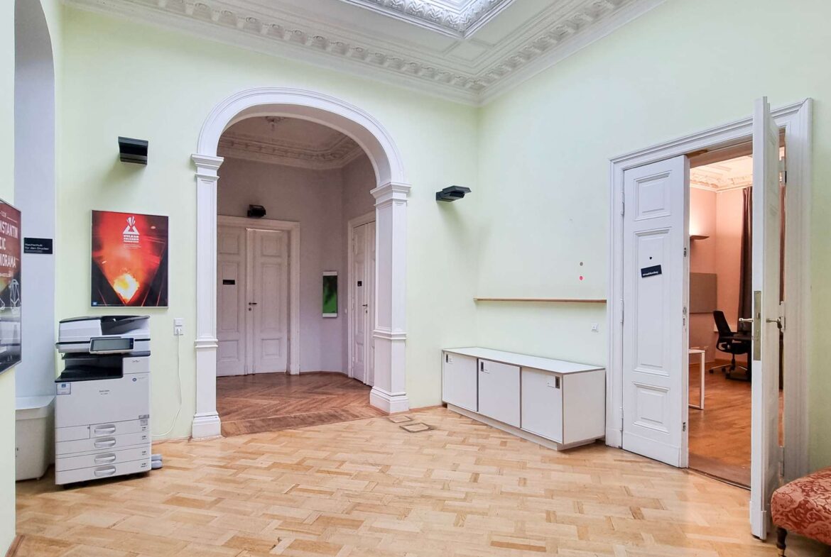 Interior hallway with pale green walls, ornate white ceiling molding, and an arched doorway leading to another room; a printer sits on a stand left.