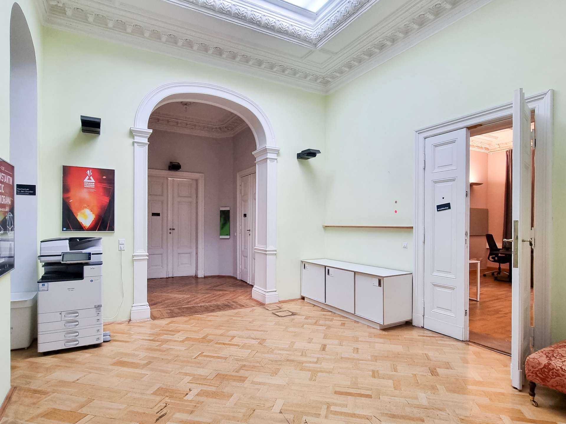 Interior hallway with pale green walls, ornate white ceiling molding, and an arched doorway leading to another room; a printer sits on a stand left.