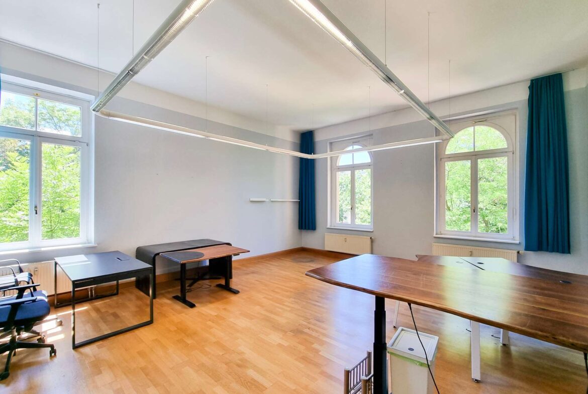 Bright classroom with wooden floor, arched windows, blue curtains, and long suspended fluorescent lights.