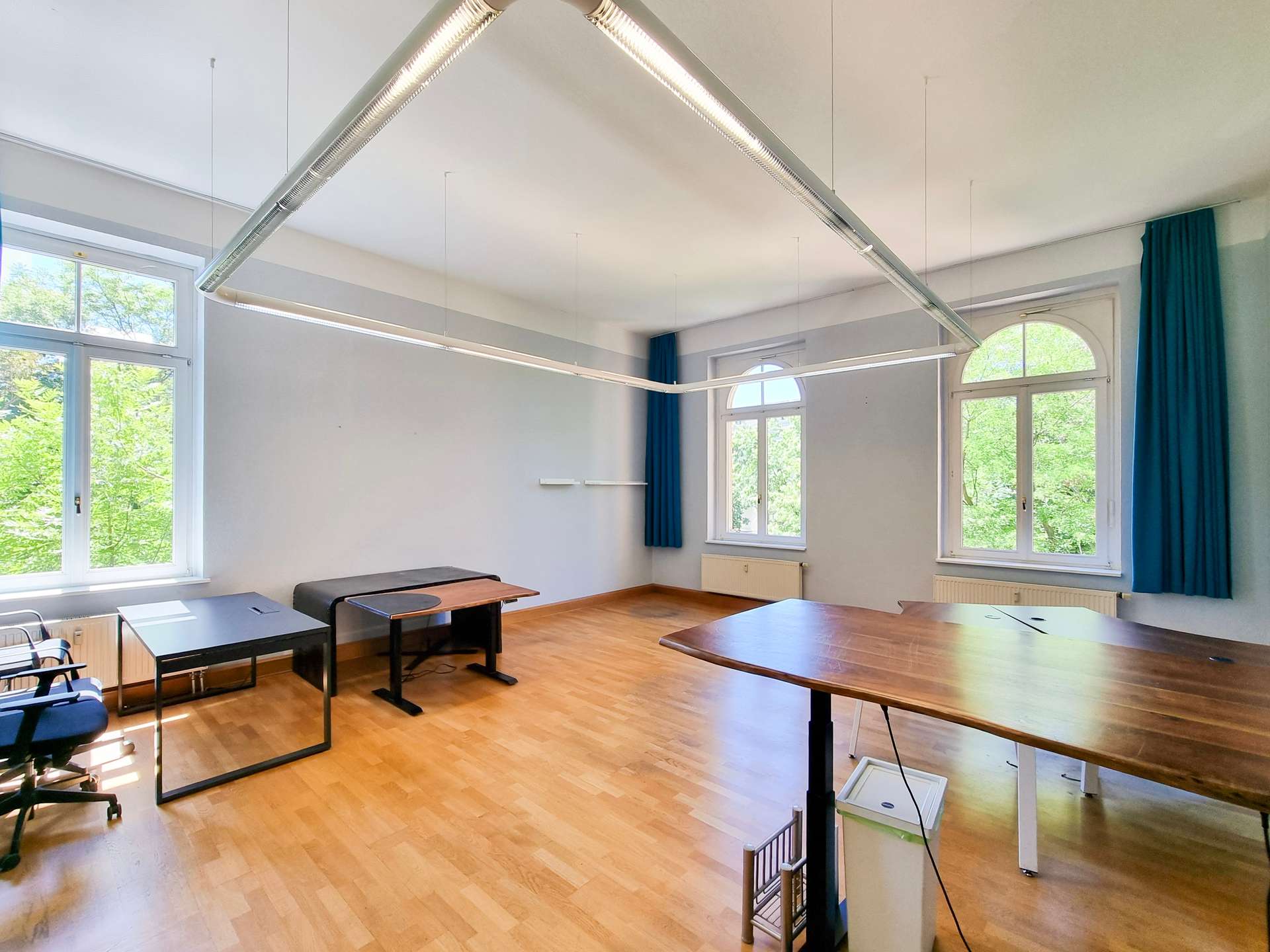 Bright classroom with wooden floor, arched windows, blue curtains, and long suspended fluorescent lights.