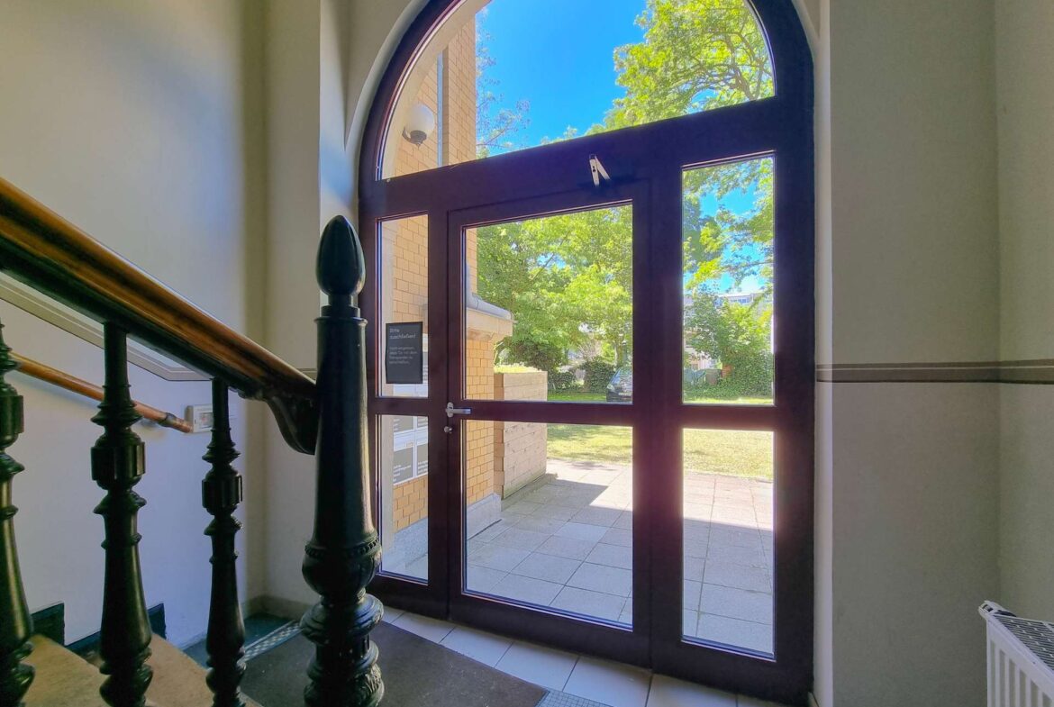 Inside building lobby with dark wood-framed glass door, arched window above, and ornate black staircase balusters nearby. Outside shows trees and brick building.