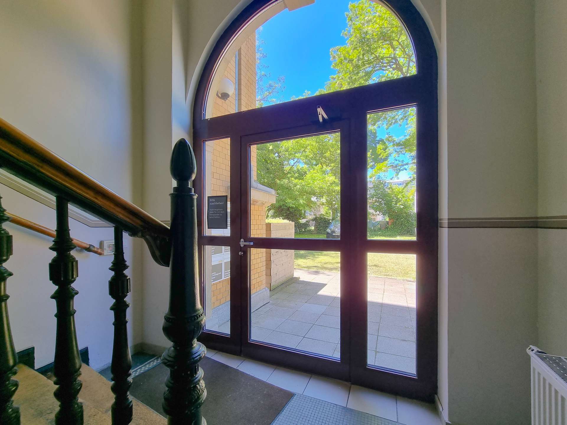 Inside building lobby with dark wood-framed glass door, arched window above, and ornate black staircase balusters nearby. Outside shows trees and brick building.