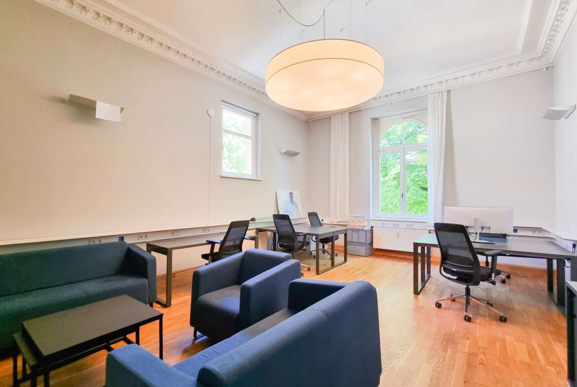 Bright conference/meeting room with blue lounge seating around black tables and laptops, a large round pendant light, and tall arched window letting in greenery.