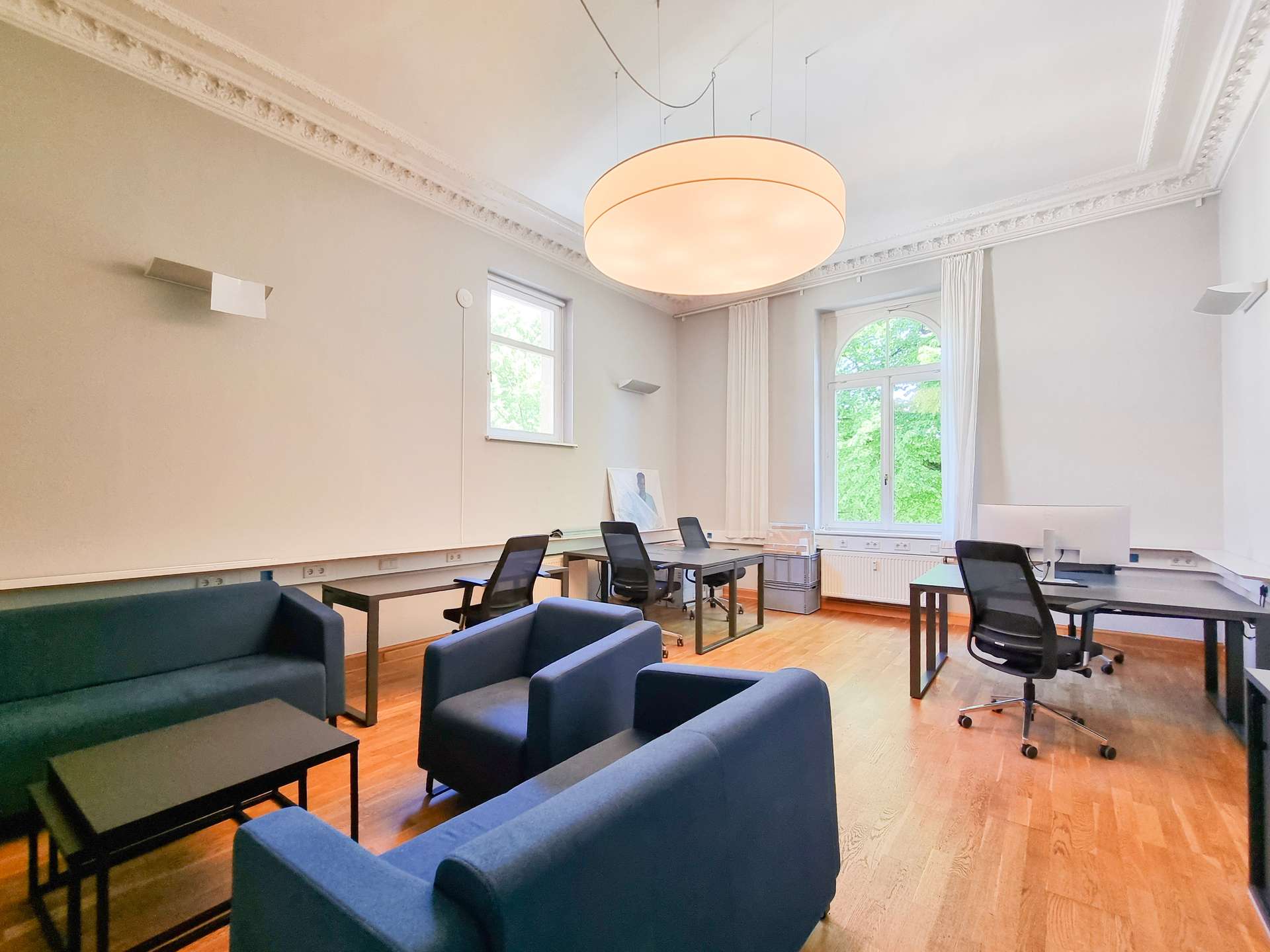 Bright conference/meeting room with blue lounge seating around black tables and laptops, a large round pendant light, and tall arched window letting in greenery.