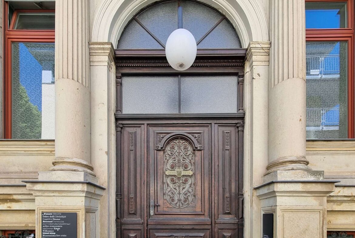 Front entrance of a historic building with a large carved wooden door, fluted columns, and a white oval light fixture above.