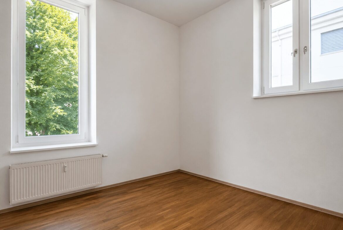 Empty white room with wooden floor, two large windows letting in daylight, radiator under the left window.