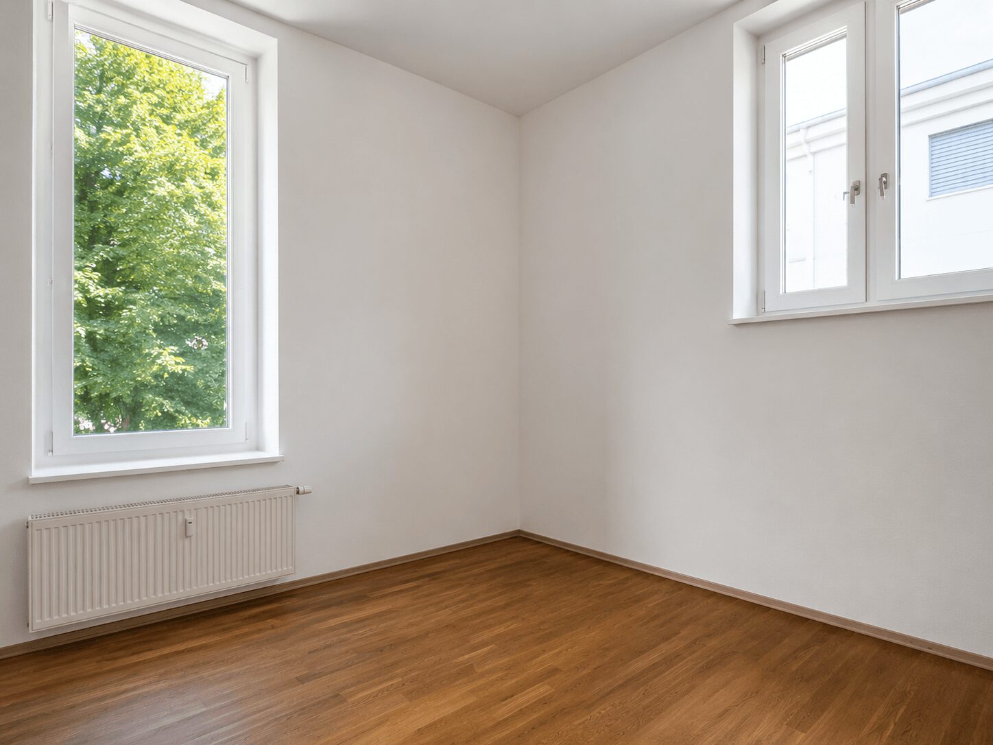 Empty white room with wooden floor, two large windows letting in daylight, radiator under the left window.