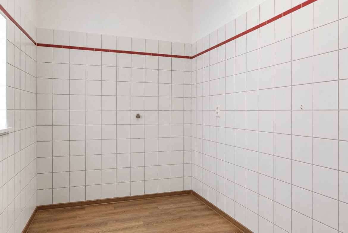 Empty tiled room with white square tiles, a red border tile near the top, and a wood-tone floor; a small pipe protrudes from the wall.