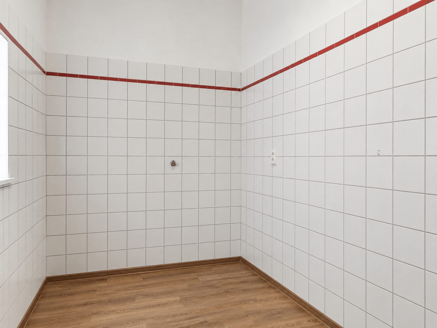 Empty tiled room with white square tiles, a red border tile near the top, and a wood-tone floor; a small pipe protrudes from the wall.