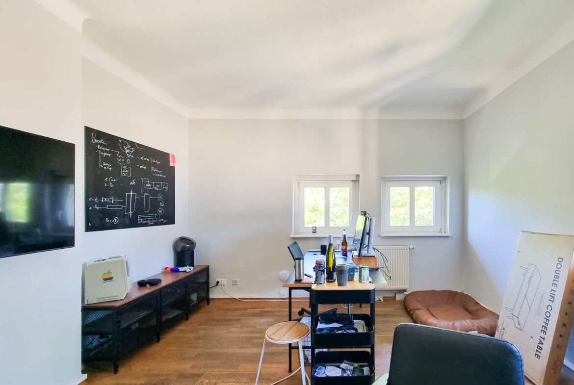Bright home office with a chalkboard wall on the left, a large flat-screen TV, and a long desk cluttered with laptops, water bottles, and cups; a cardboard box leans against the wall on the right side of the room.