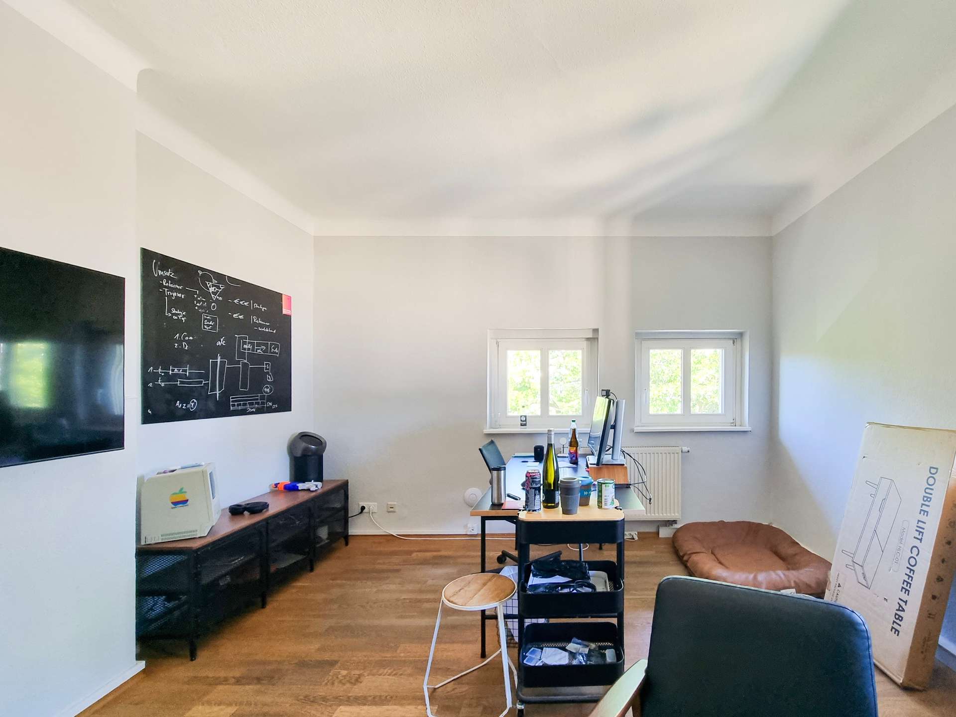 Bright home office with a chalkboard wall on the left, a large flat-screen TV, and a long desk cluttered with laptops, water bottles, and cups; a cardboard box leans against the wall on the right side of the room.