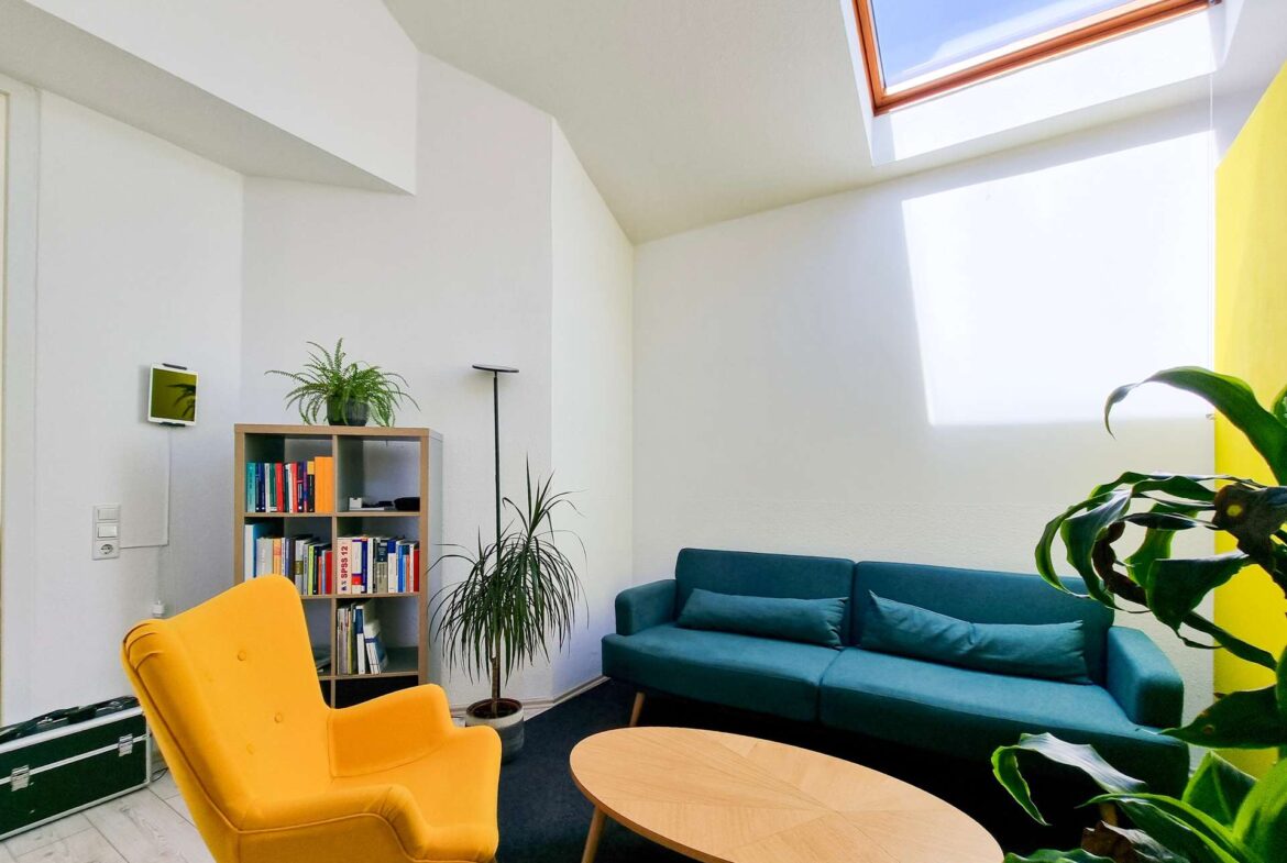 Bright living room with a yellow armchair, teal sofa, round wooden coffee tables, bookshelf, and tall plants under a skylight.
