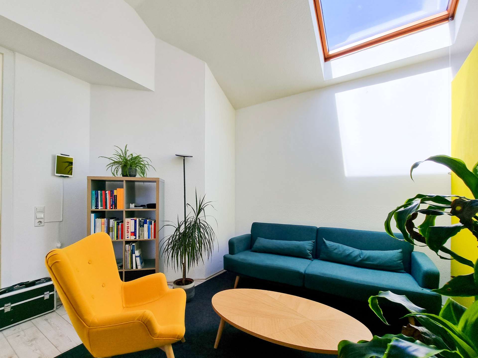 Bright living room with a yellow armchair, teal sofa, round wooden coffee tables, bookshelf, and tall plants under a skylight.