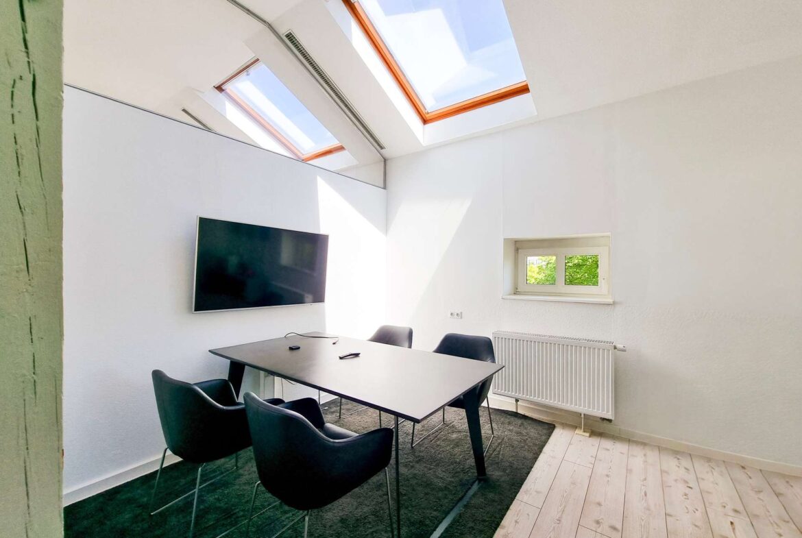 Modern meeting room with a rectangular table, black chairs, a wall-mounted TV, skylight windows, and a radiator beneath a small window.