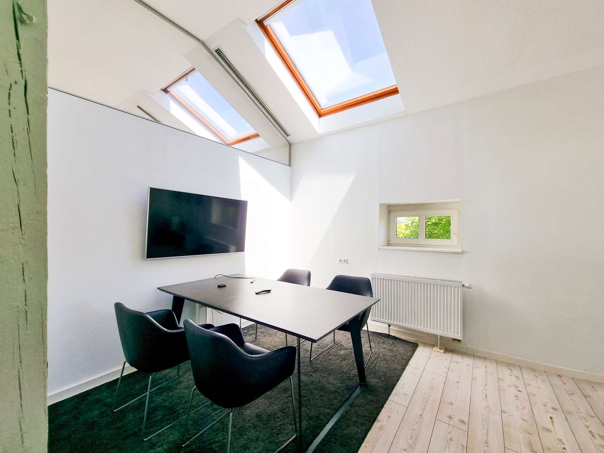 Modern meeting room with a rectangular table, black chairs, a wall-mounted TV, skylight windows, and a radiator beneath a small window.