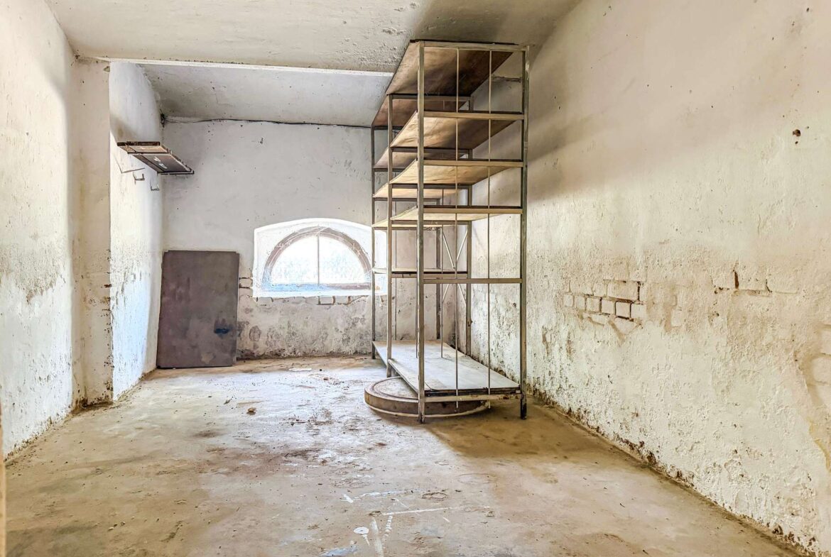 Abandoned industrial room with tall metal shelving unit and an arched window in the back wall, dusty concrete floor and weathered white walls.