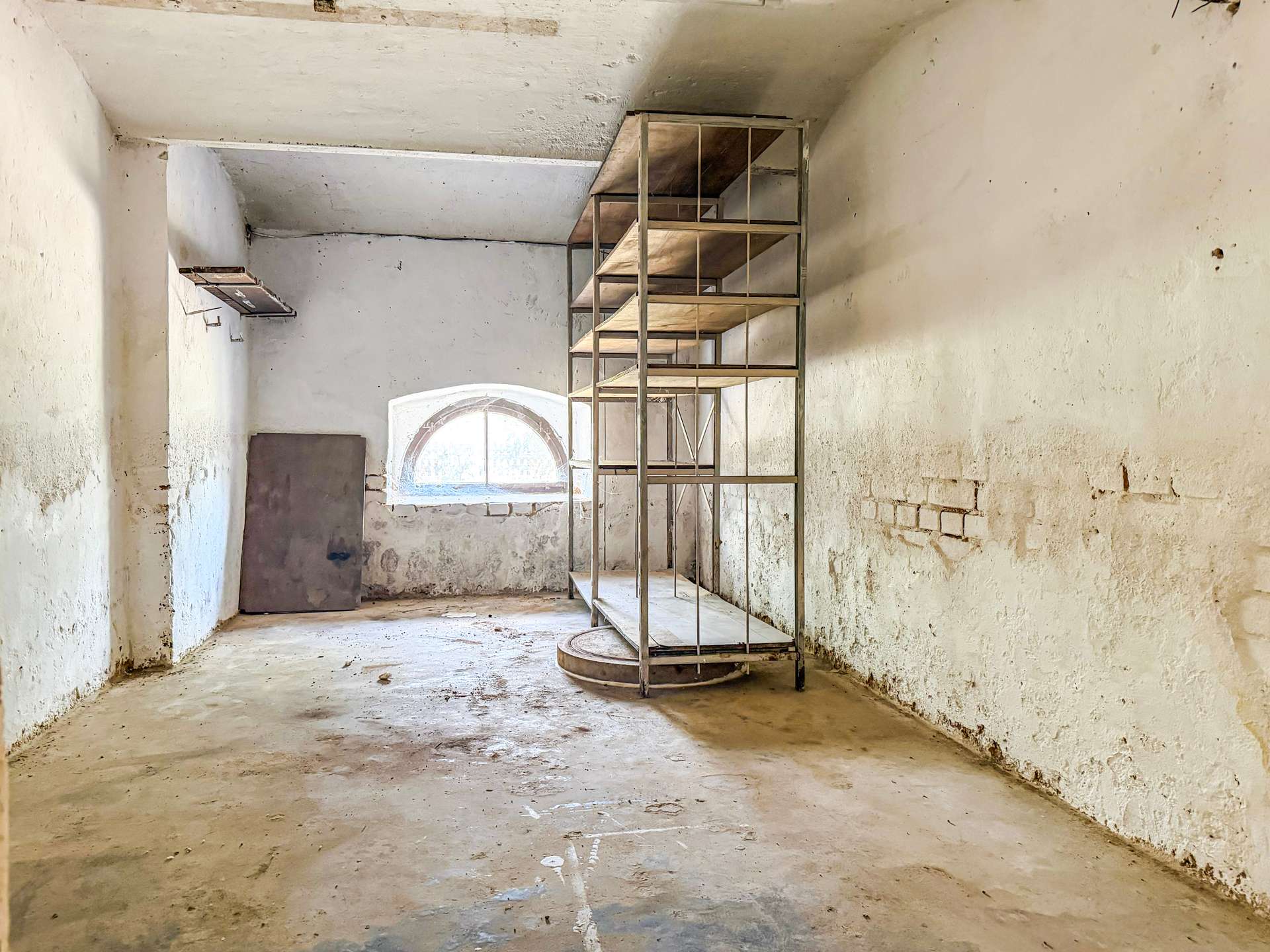 Abandoned industrial room with tall metal shelving unit and an arched window in the back wall, dusty concrete floor and weathered white walls.