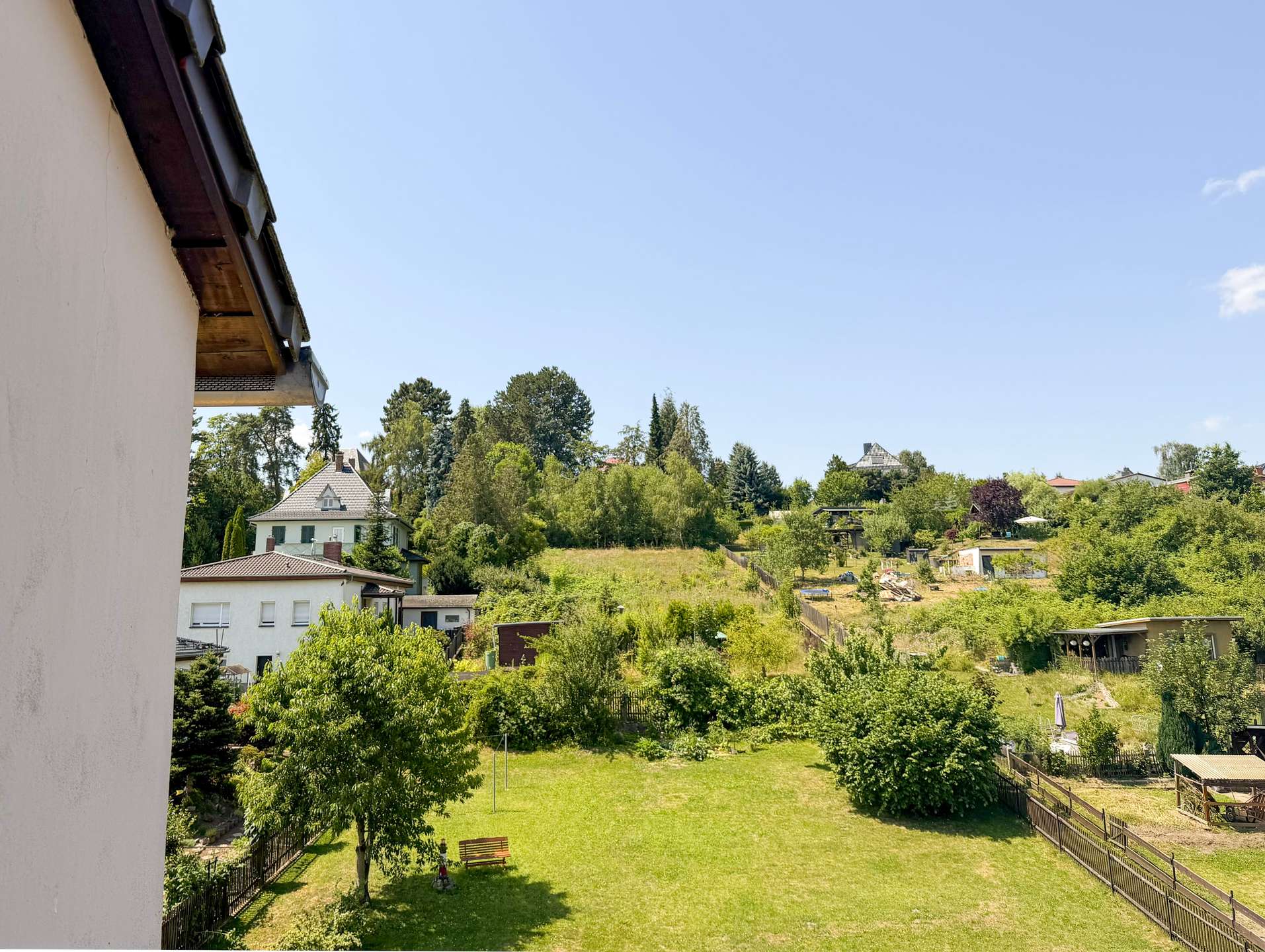 Backyard with a green lawn, trees, and a wooden fence; houses in the distance under a clear blue sky.