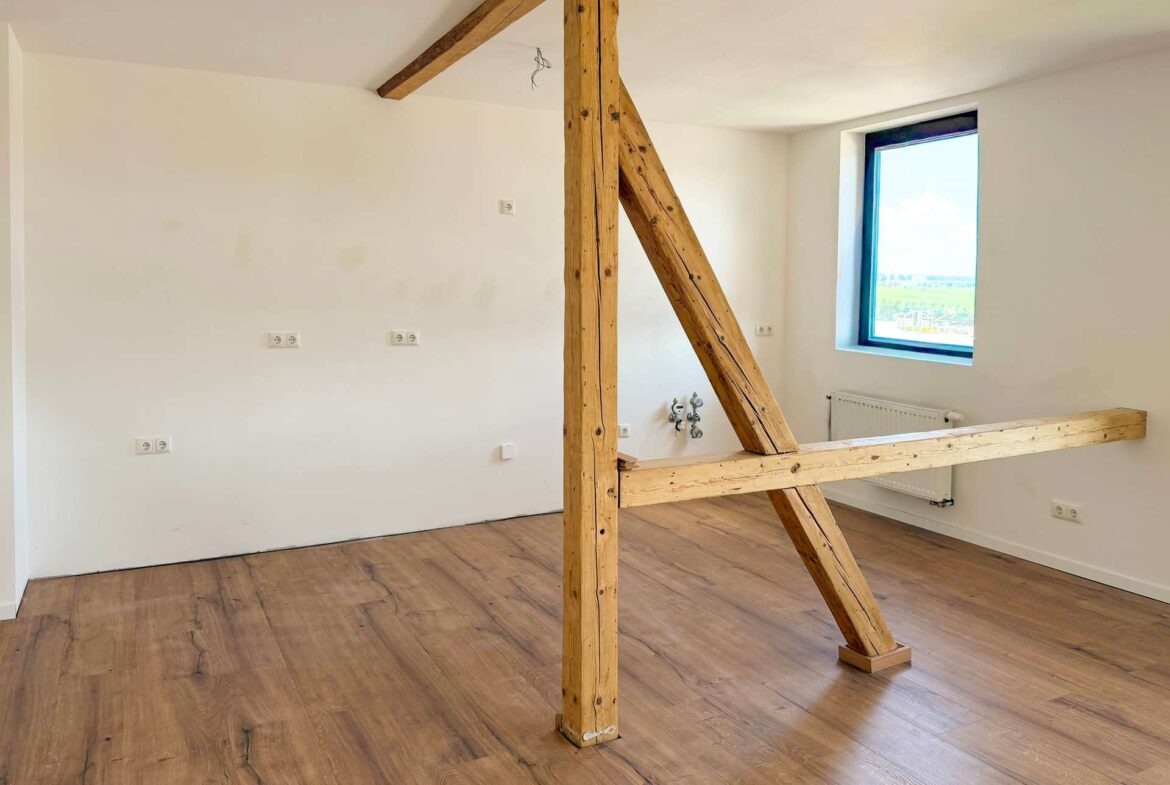 Interior of a bright room with exposed wooden support beams and a diagonal brace, white walls, and wood-look flooring.