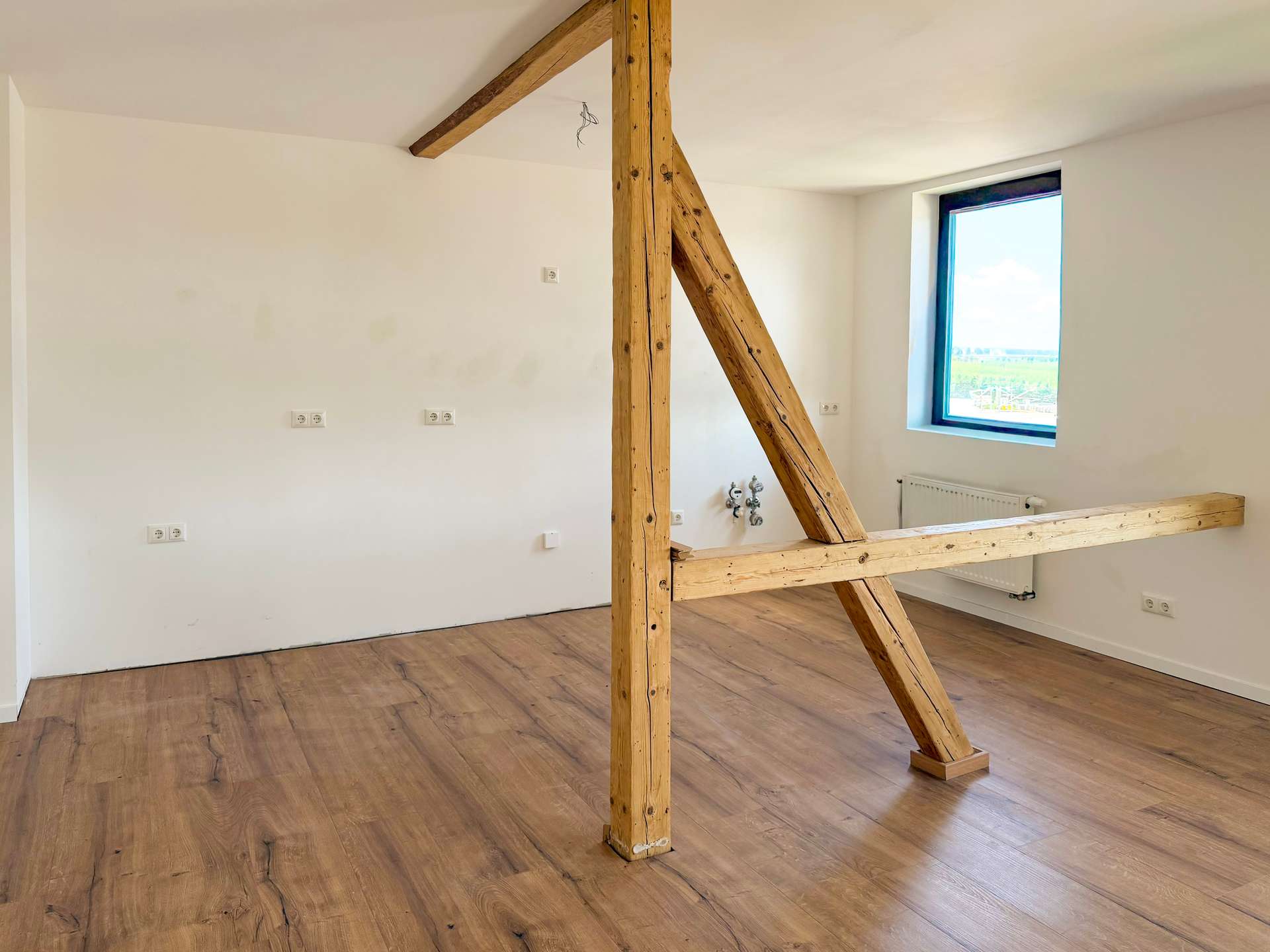 Interior of a bright room with exposed wooden support beams and a diagonal brace, white walls, and wood-look flooring.