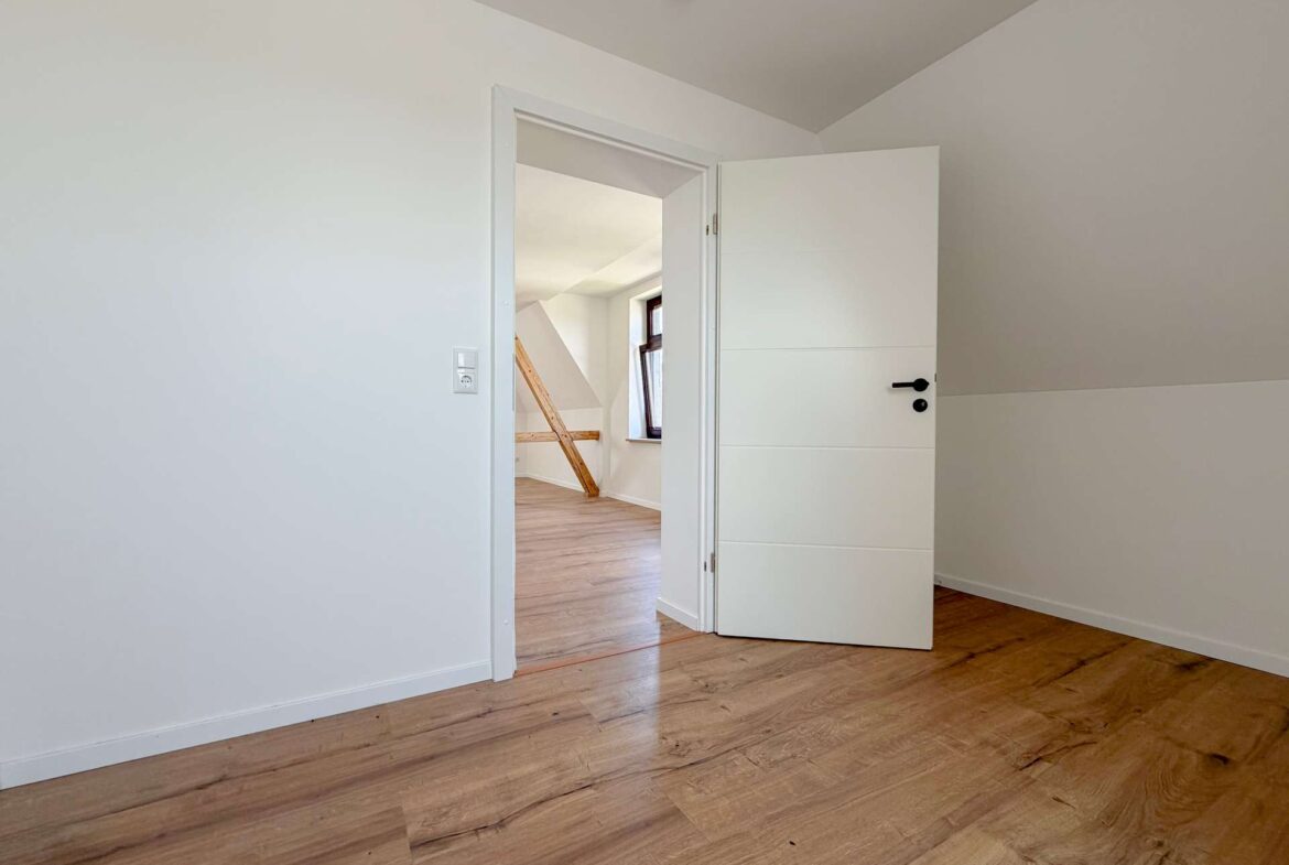 Empty white-walled room with light wooden floor, open door to a second room showing a diagonal beam and window outside the doorway.