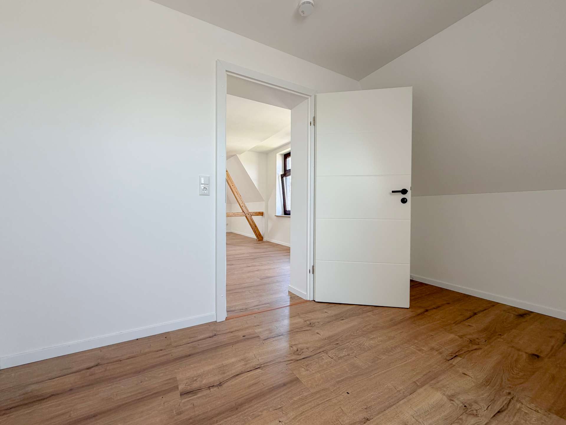 Empty white-walled room with light wooden floor, open door to a second room showing a diagonal beam and window outside the doorway.