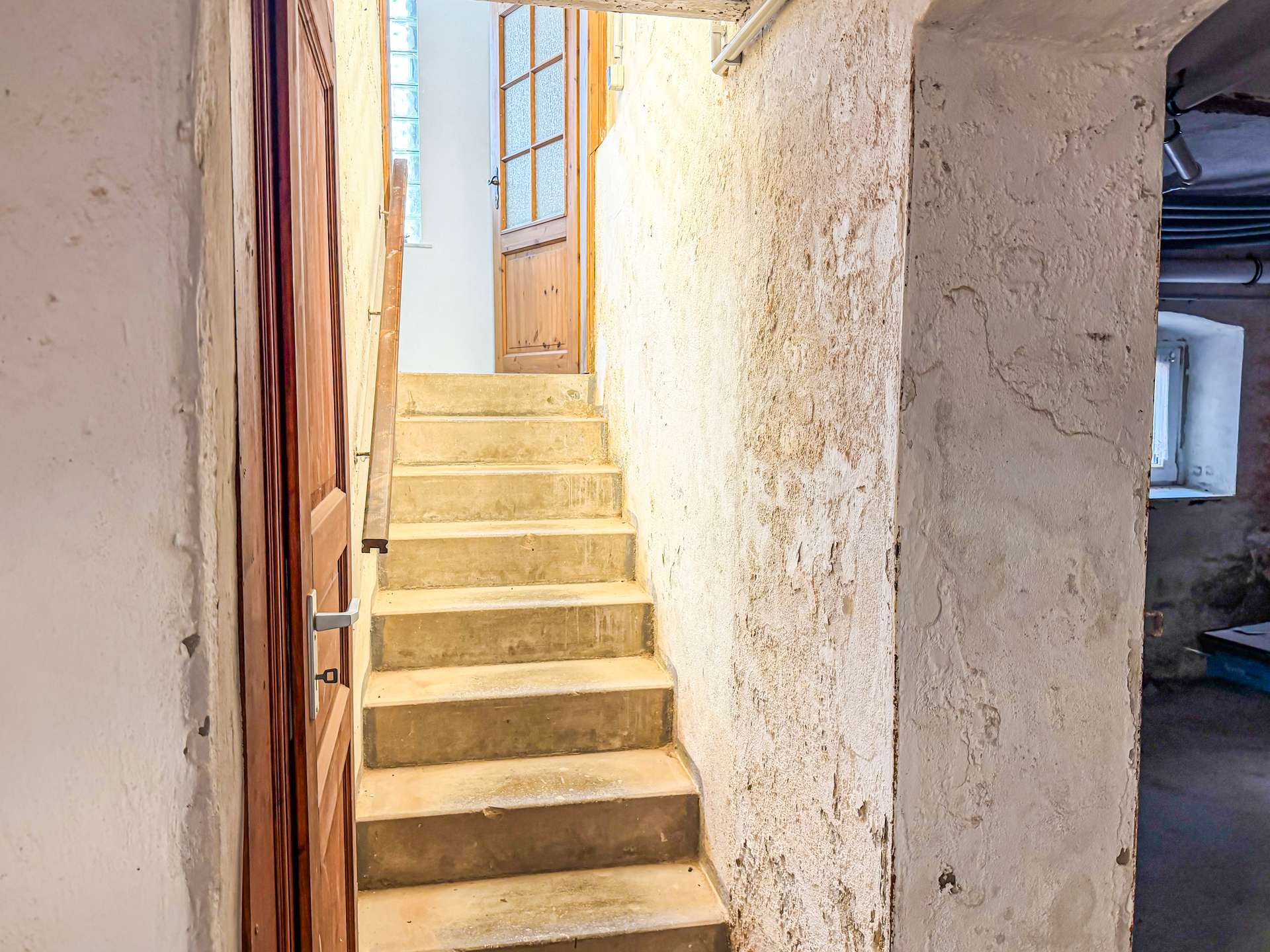Narrow basement stairwell with concrete steps and a wooden door on the left, textured white walls surrounding the stairs.