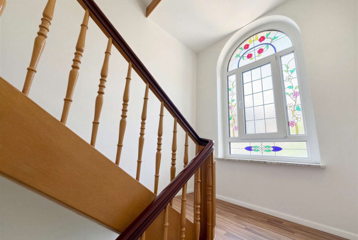 Wooden staircase with turned balusters and a dark handrail beside a large arched stained-glass window.